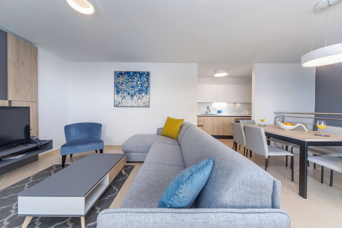 Ein modernes Wohnzimmer mit Sofa, Fernseher, Esstisch und einer Küche im Hintergrund.