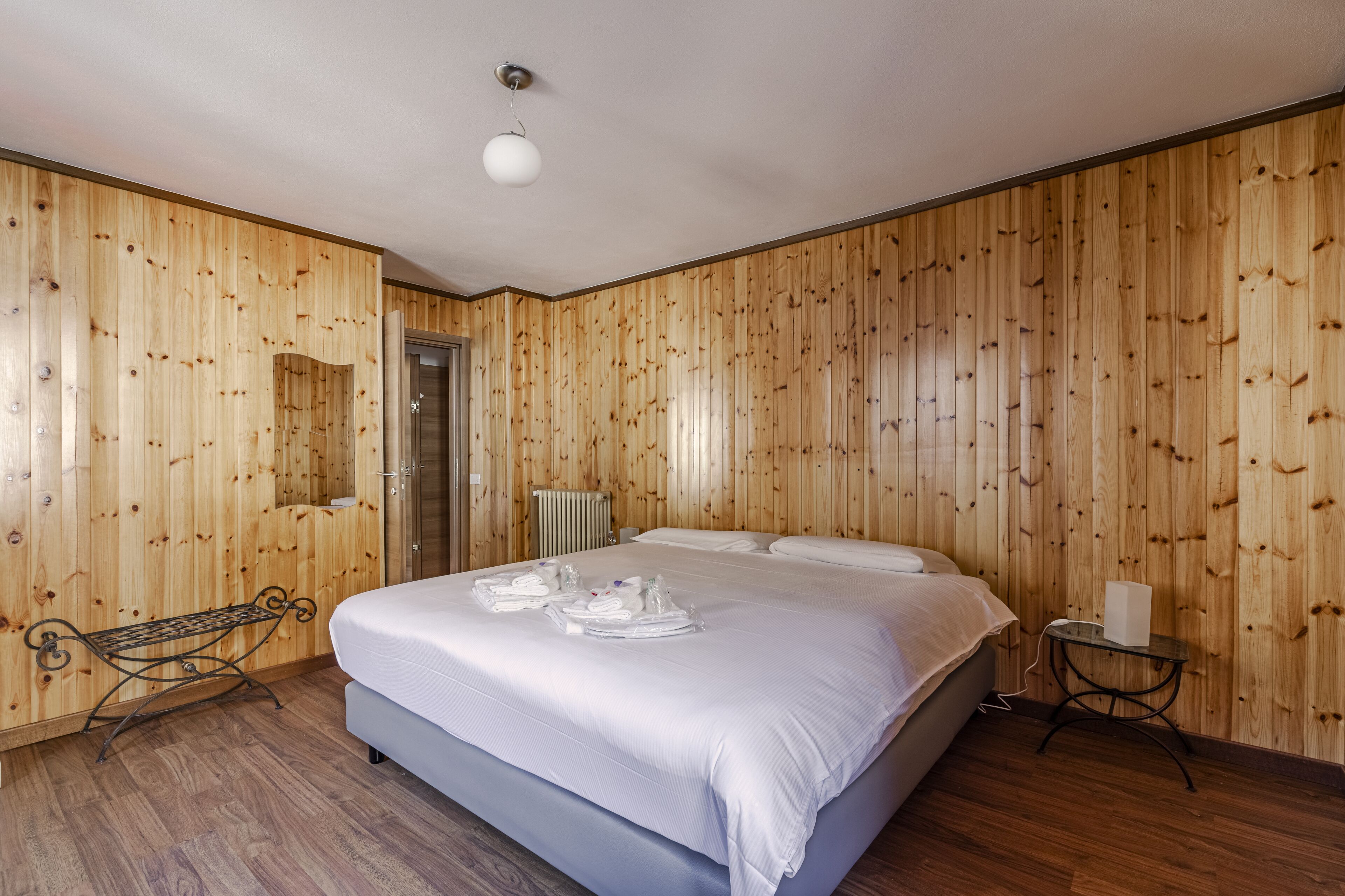 Una habitación con paneles de madera, una cama, un banco y una pequeña mesa con una lámpara.