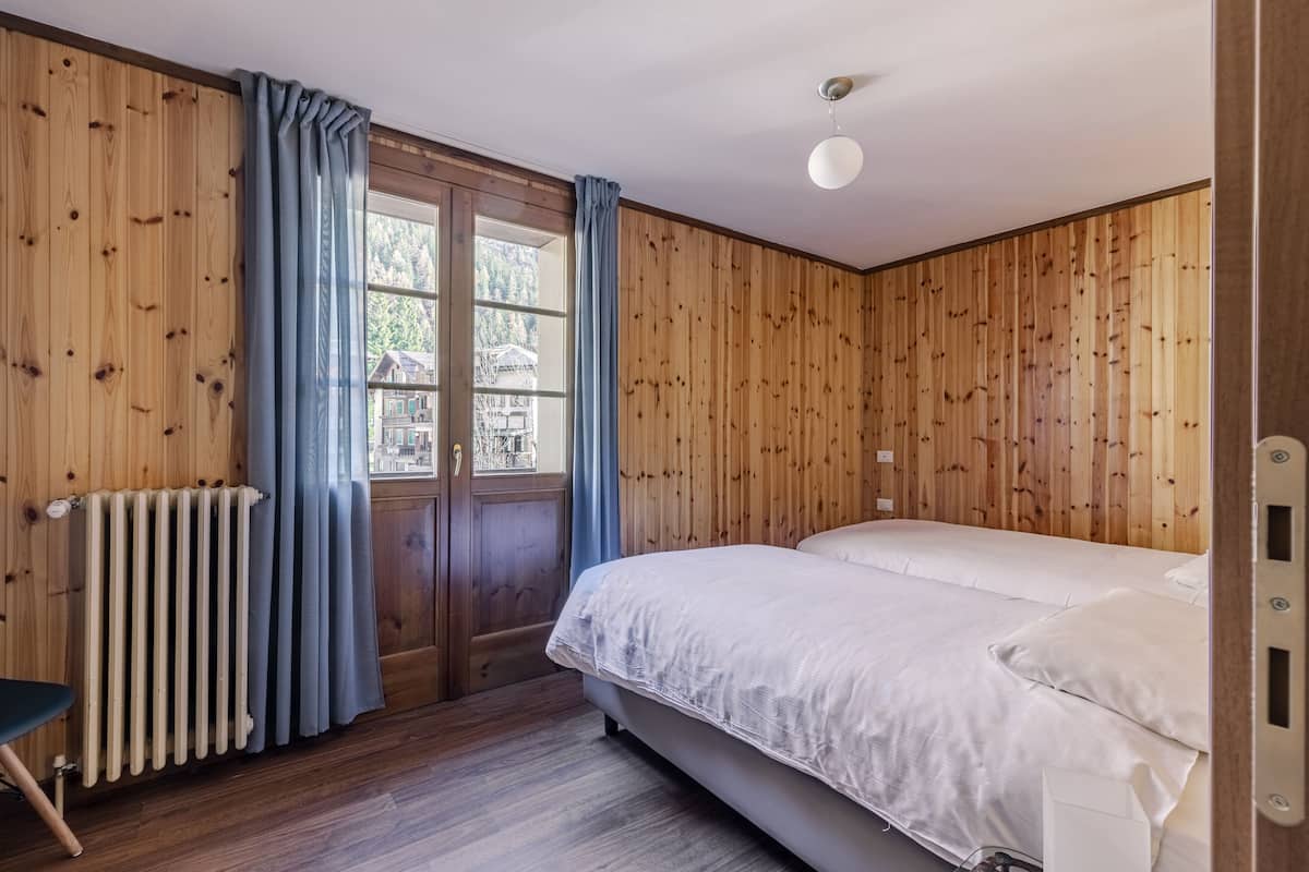 Une pièce aux murs en bois, un lit avec une literie blanche, un radiateur, une fenêtre avec des rideaux et une vue sur un bâtiment à l’extérieur.
