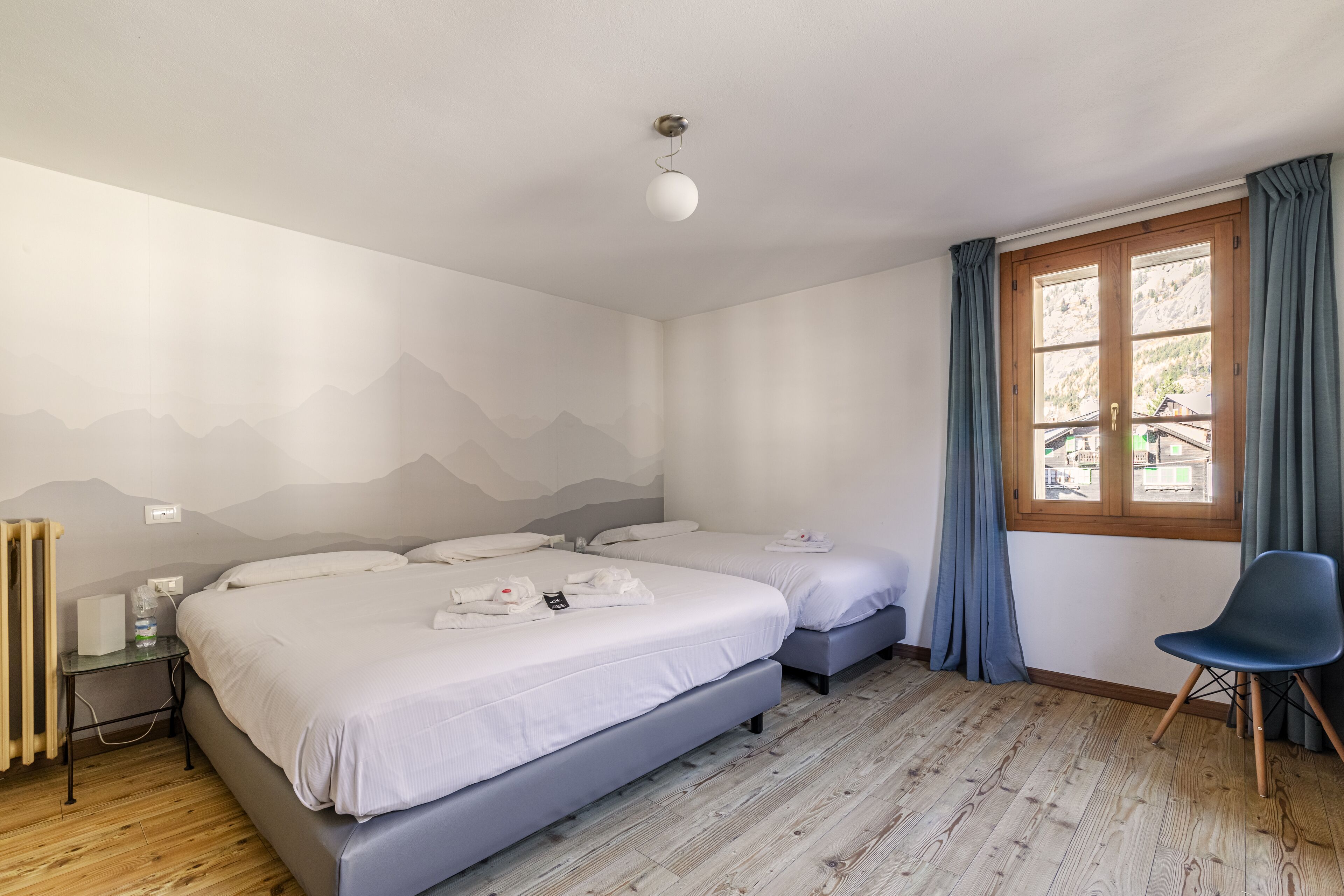 A hotel room with two beds, a wooden floor, a radiator, a window with curtains, and a chair.