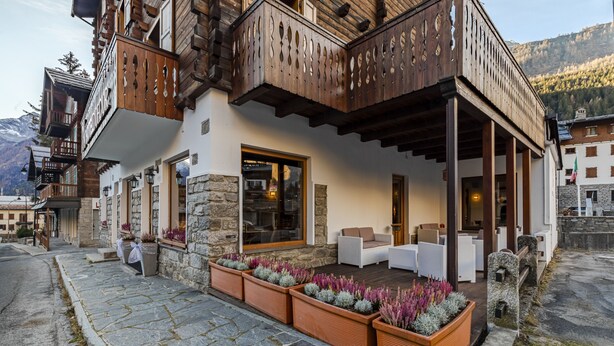 A hotel with a stone facade, wooden balconies, outdoor seating, and a mountain backdrop. - Hotel Dufour (Macugnaga)