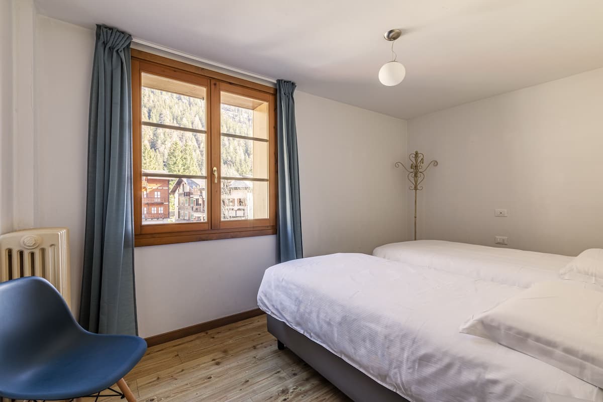 Une chambre à coucher comprenant un lit, une chaise, un radiateur, une fenêtre avec des rideaux et une vue sur les bâtiments à l’extérieur.