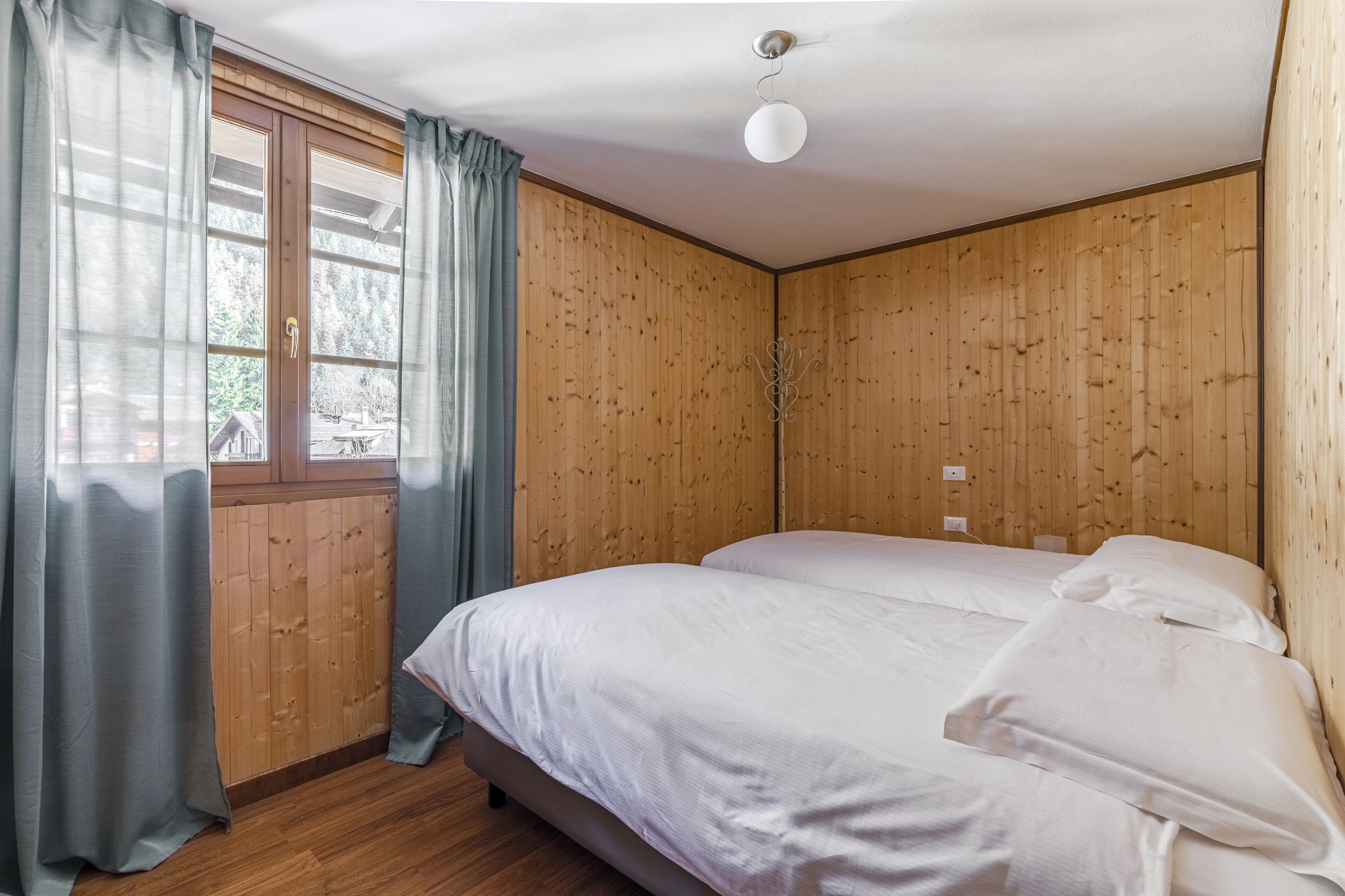 Una habitación con paneles de madera, una cama con ropa de cama blanca, una ventana con cortinas y una lámpara de techo.