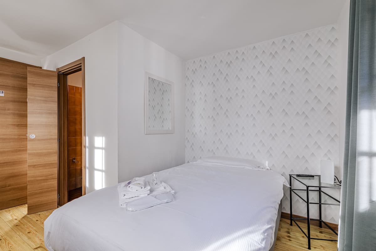 Une chambre d’hôtel avec une armoire en bois, un lit avec du linge de maison blanc, une petite table avec une télécommande et un mur à motifs.