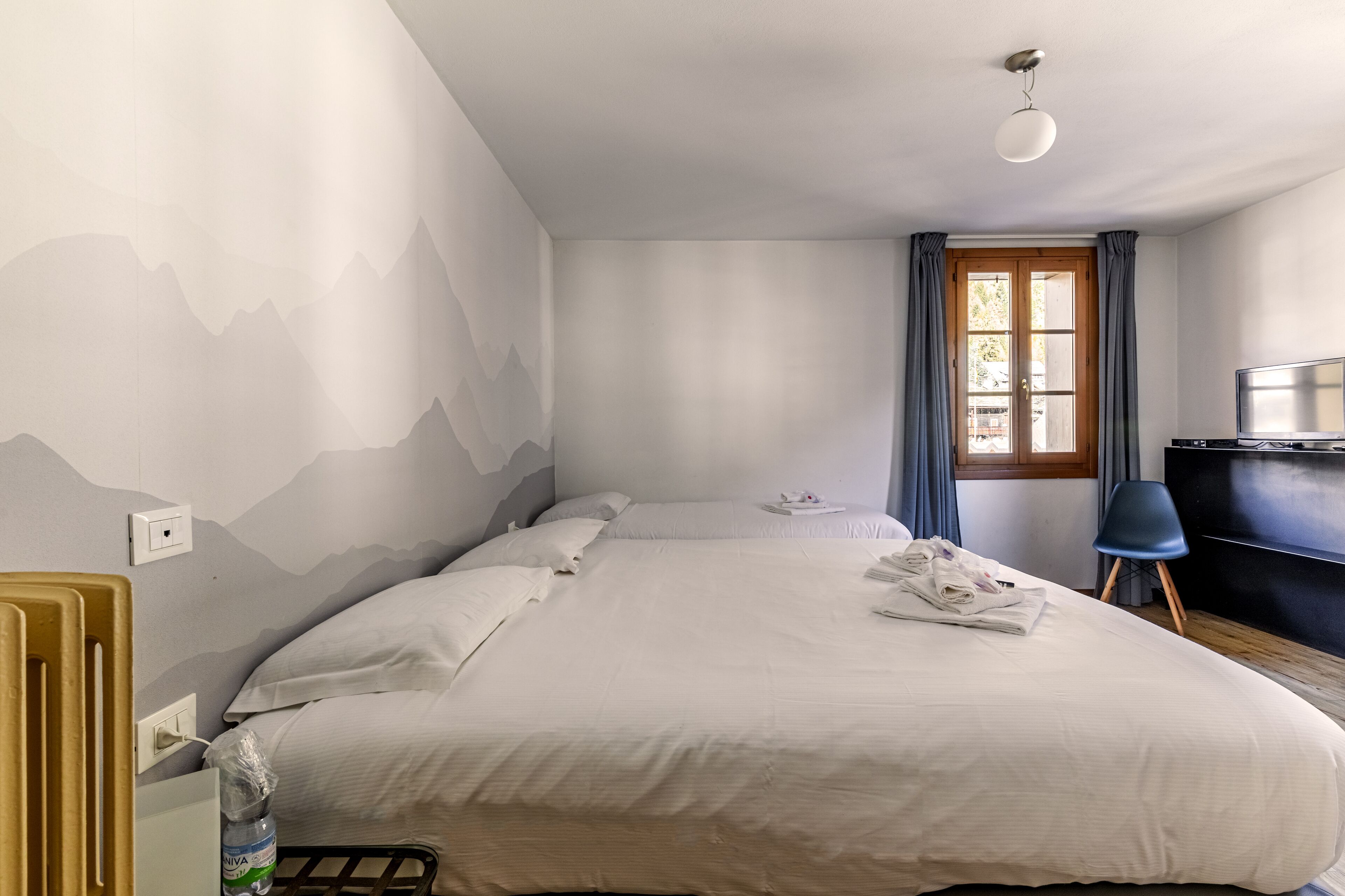Een net opgemaakt bed met witte lakens, een houten hoofdbord, een klein nachtkastje en een blauwe stoel.