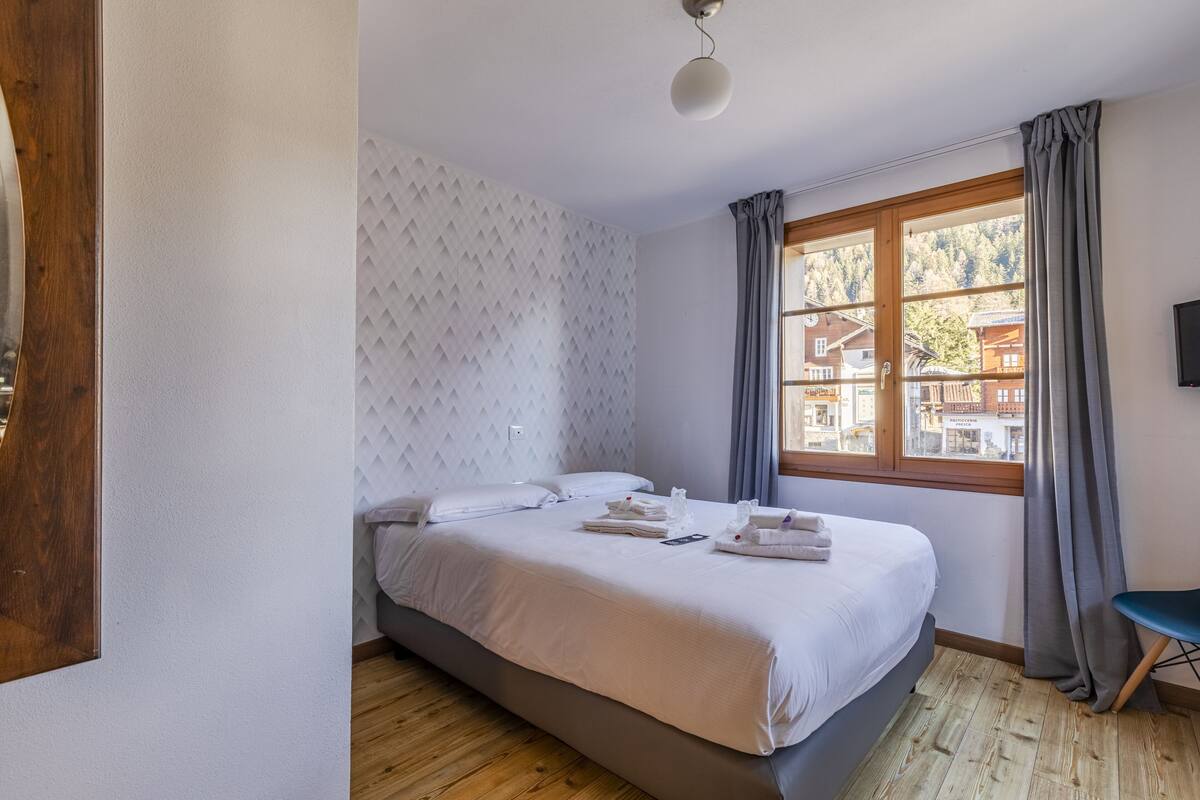Un lit bien fait, avec du linge de lit et des serviettes blanches, un plancher en bois, une fenêtre avec des rideaux et un téléviseur fixé au mur.
