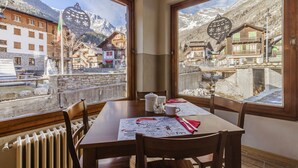 A dining area with a table set for four, large windows offering a view of a mountain village with traditional buildings and a stone wall. - Hotel Dufour (Macugnaga)