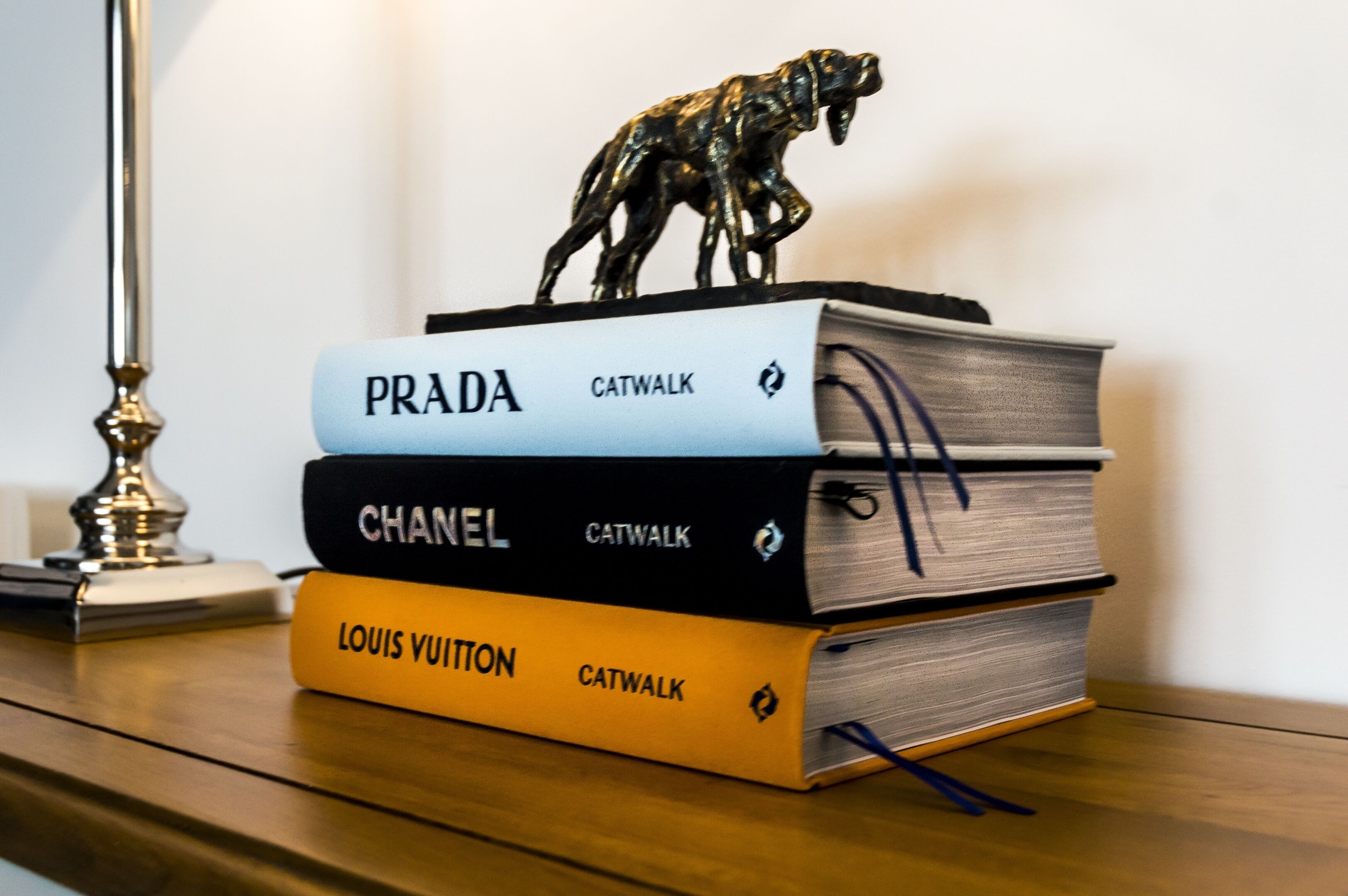 Une pile de livres dont les titres sont « Prada », « Chanel » et « Louis Vuitton », posée sur une surface en bois.