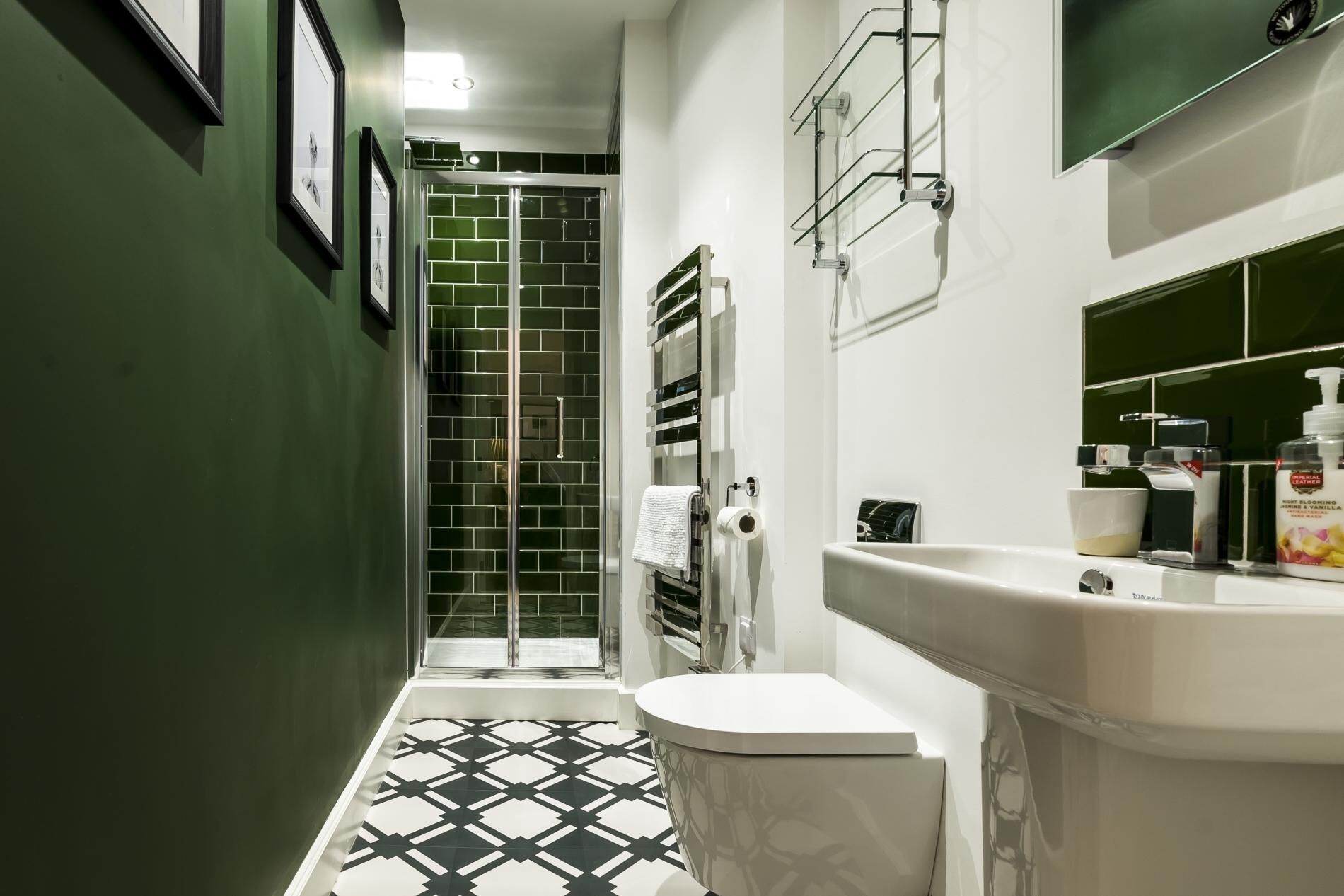 A narrow bathroom with a black and white patterned floor, a white toilet, a glass shower enclosure, and a white sink with a mirror above it.