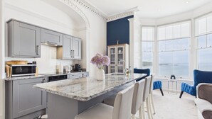 Een moderne keuken met een marmeren eiland, grijze keukenkastjes en uitzicht op zee door grote ramen.