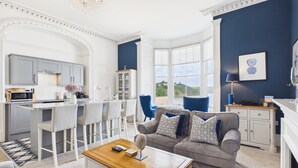 Een ruime woonkamer met een grijze bank, witte stoelen, een houten salontafel en uitzicht op zee door grote ramen.