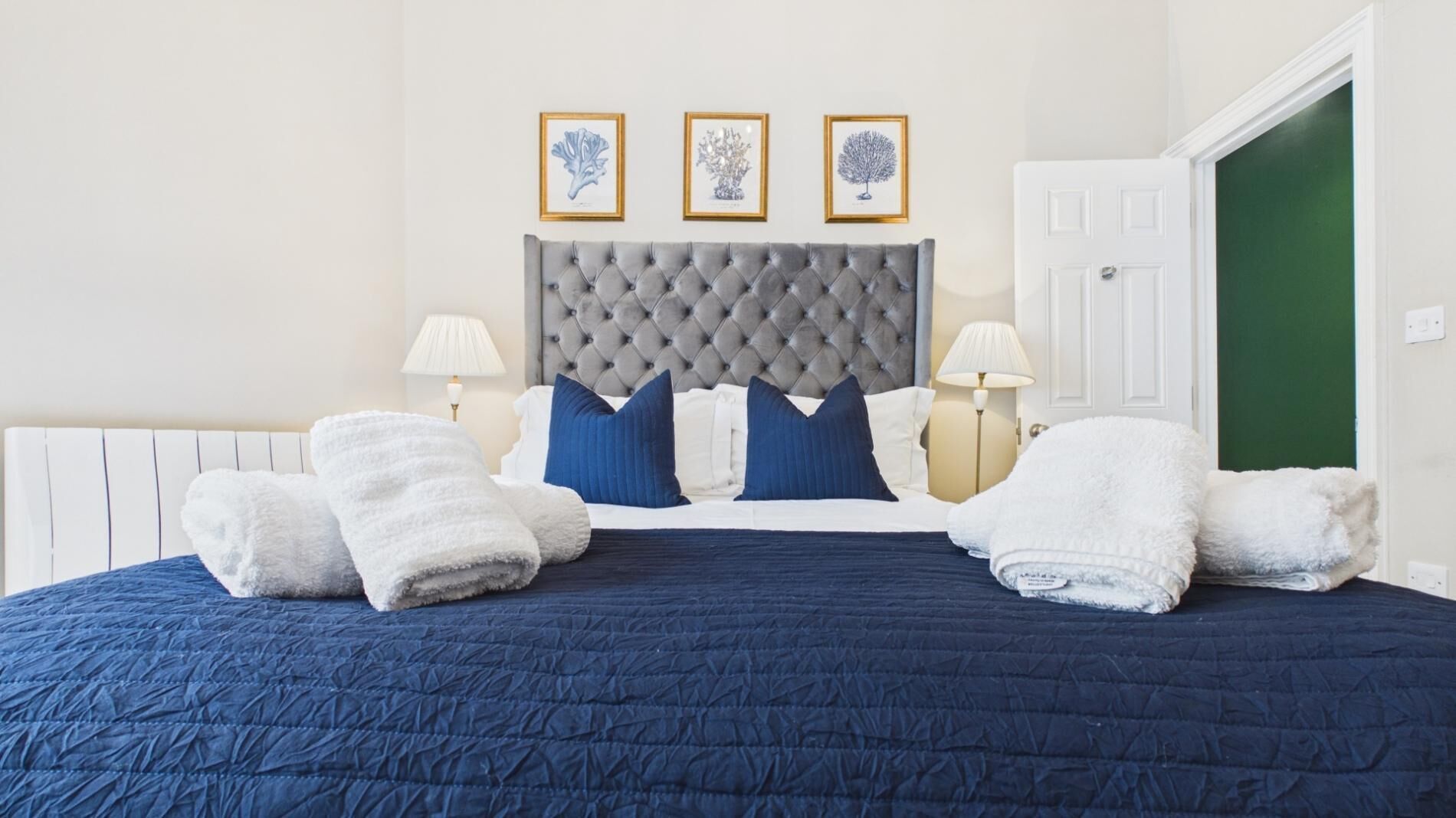 A neatly made bed with a blue quilt, grey headboard, and white towels. There are three framed pictures on the wall above the bed.