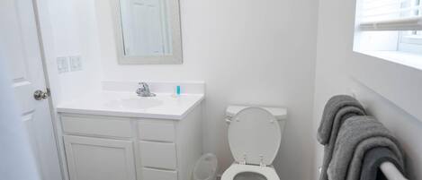 Un bagno piccolo e pulito, con sanitari bianchi, uno specchio sopra il lavandino e una finestra con persiane.