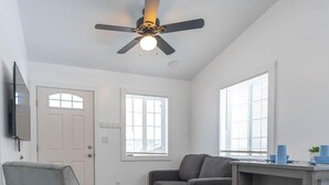A living room with a ceiling fan, a gray sofa, a small table with blue cups, and a TV mounted on the wall.