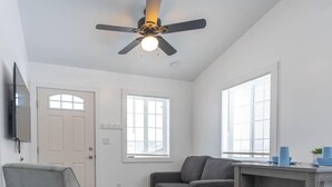 A living room with a ceiling fan, a gray sofa, a small table with blue cups, and a TV mounted on the wall.