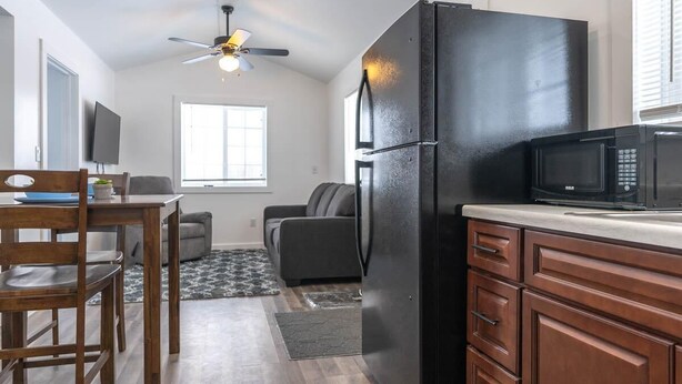 A compact kitchen with a black refrigerator, a microwave, wooden cabinets, a ceiling fan, and a dining area with a table and chairs. - Sparkling Clean! 2 Bedroom Cottage- Located in Beautiful Keweenaw (Calumet)