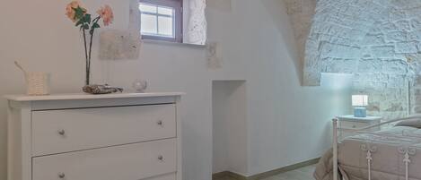 A bedroom with a white dresser, a bed with a beige cover, a small window, and a stone wall.