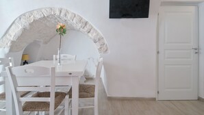 Un comedor blanco con un arco de piedra, una mesa blanca y sillas. Hay un televisor montado en la pared y una puerta blanca.