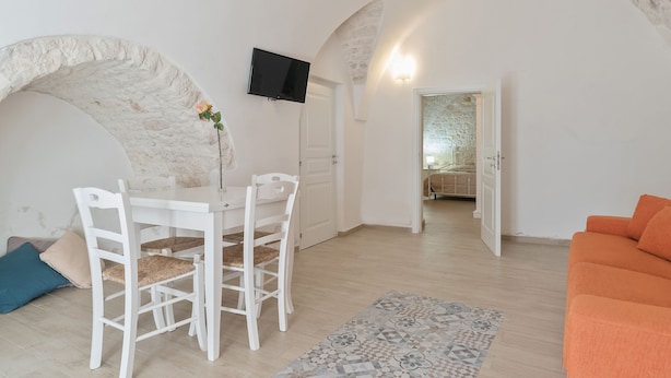 Una sala de estar con una mesa de comedor blanca, sillas blancas, un televisor montado en la pared y un sofá naranja.