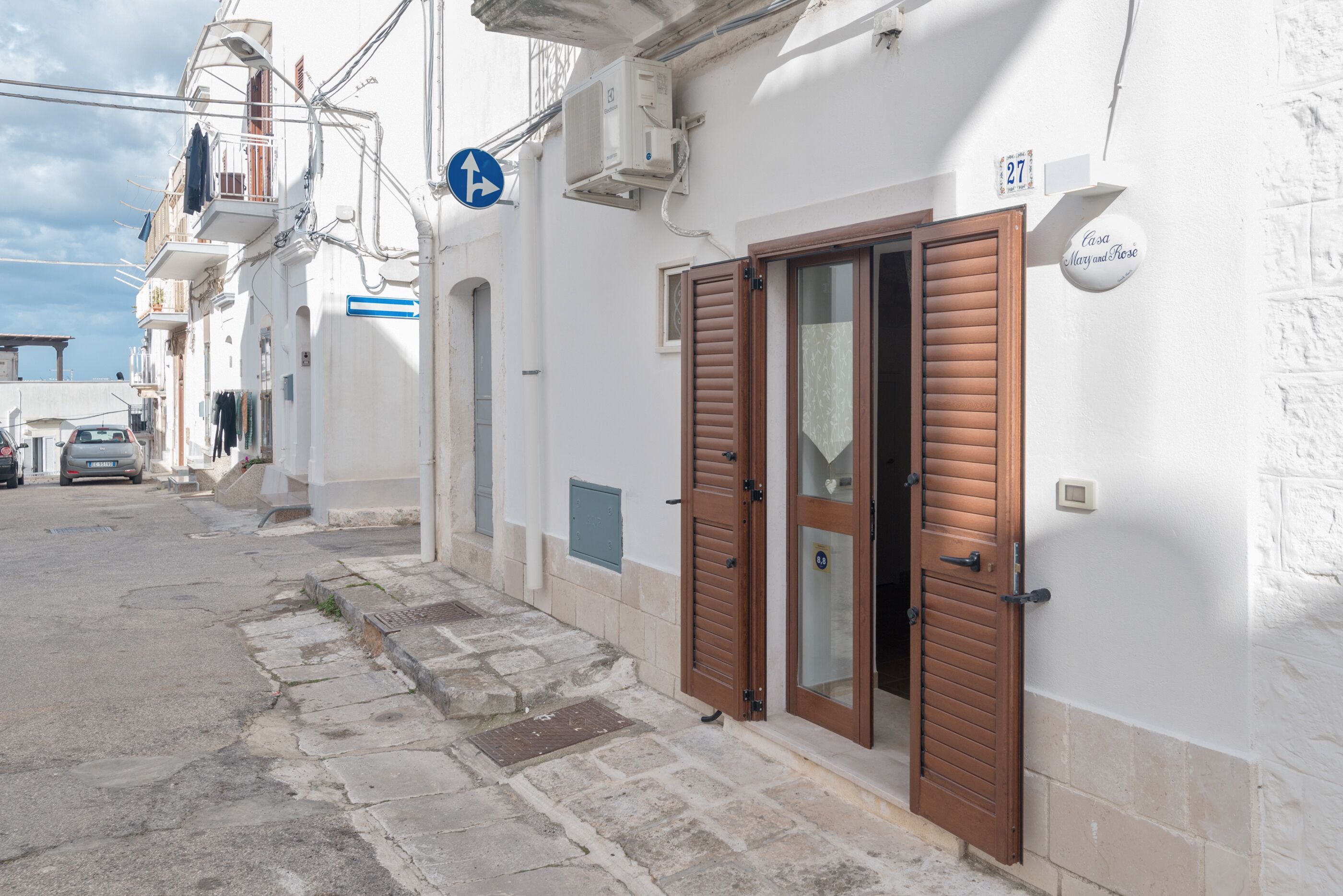 A narrow street with white buildings, a blue sign, and a house number 27.