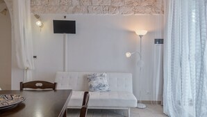 Een kamer met een witte bank, een eettafel met stoelen, een flatscreen televisie aan de wand en een vloerlamp.