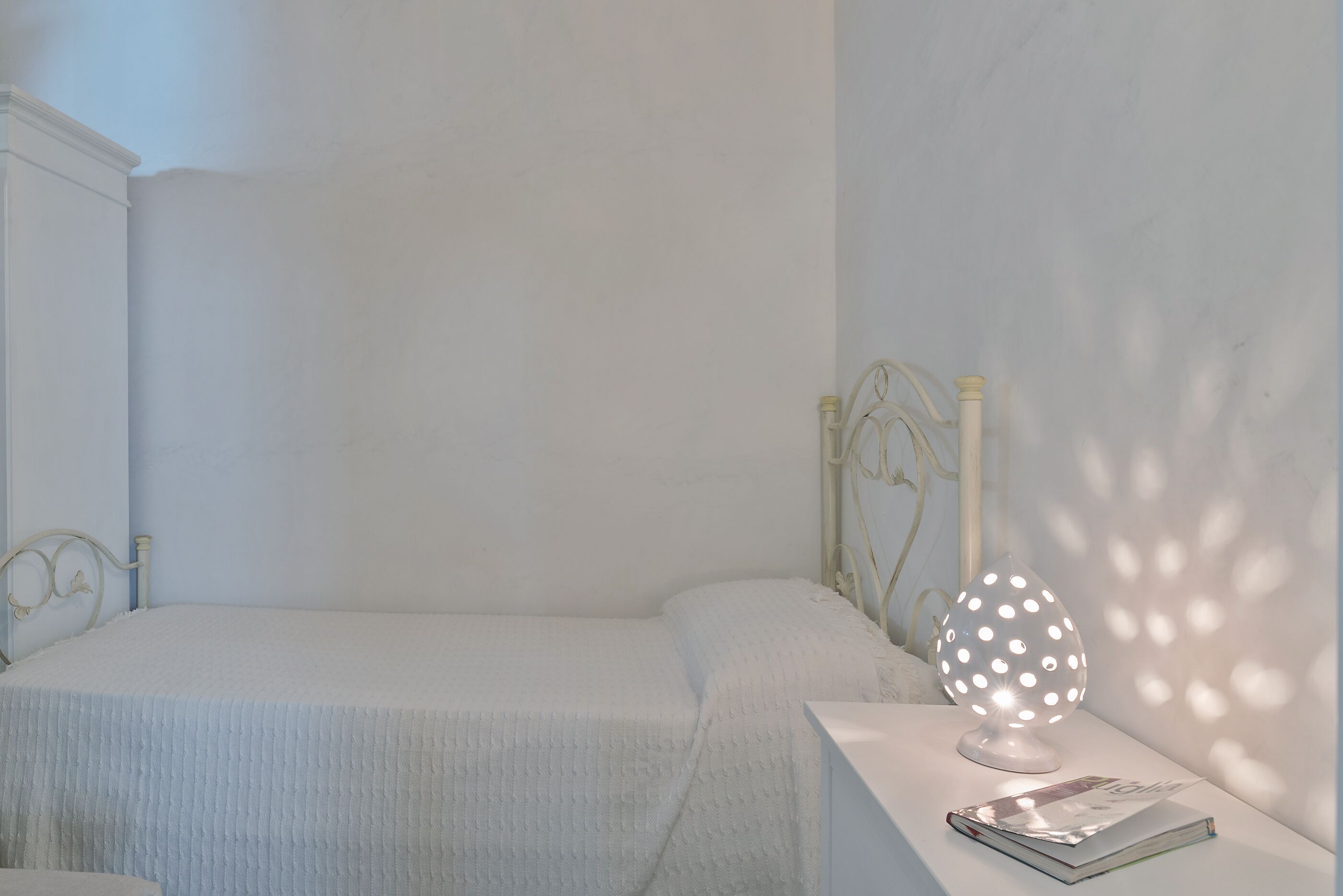 A neatly made bed with a white headboard, a bedside table with a book, and a wall-mounted lamp.