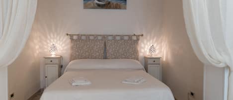 A neatly made bed with a white bedspread and two folded towels, flanked by two bedside tables with lamps, in a room with white curtains and a painting on the wall.