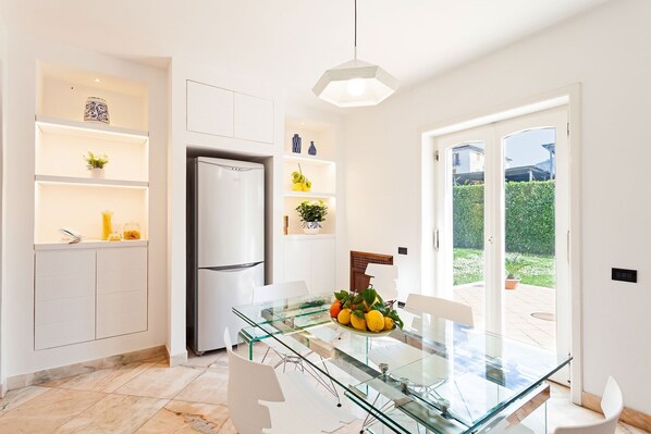 Una cocina moderna con muebles blancos, un refrigerador empotrado y una mesa de comedor de vidrio con un cuenco de fruta.