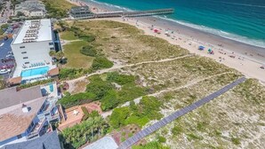 An aerial view of a coastal area with a beach, a pier, and buildings including a hotel with a swimming pool.