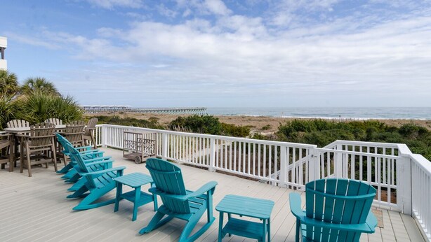 A deck with turquoise Adirondack chairs and a white railing overlooking a beach and ocean.