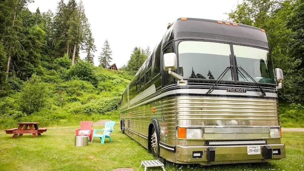 A silver bus with the brand name 'PREVOST' parked in a grassy area with picnic tables and chairs.