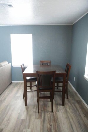 A dining area with a wooden table and four chairs, a light blue wall, and a window providing natural light.