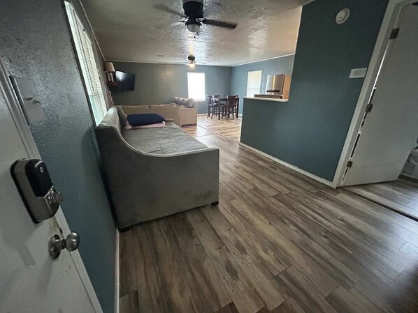 A room with a grey sofa, a dining table with chairs, a ceiling fan, and a TV mounted on the wall.