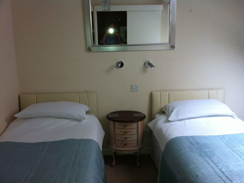 A hotel room with two single beds, a small wooden nightstand, a mirror, and a light switch on the wall.
