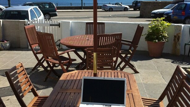 A patio with a table and chairs, a laptop, and a view of the sea and parked cars. - The Stanley (Penzance)