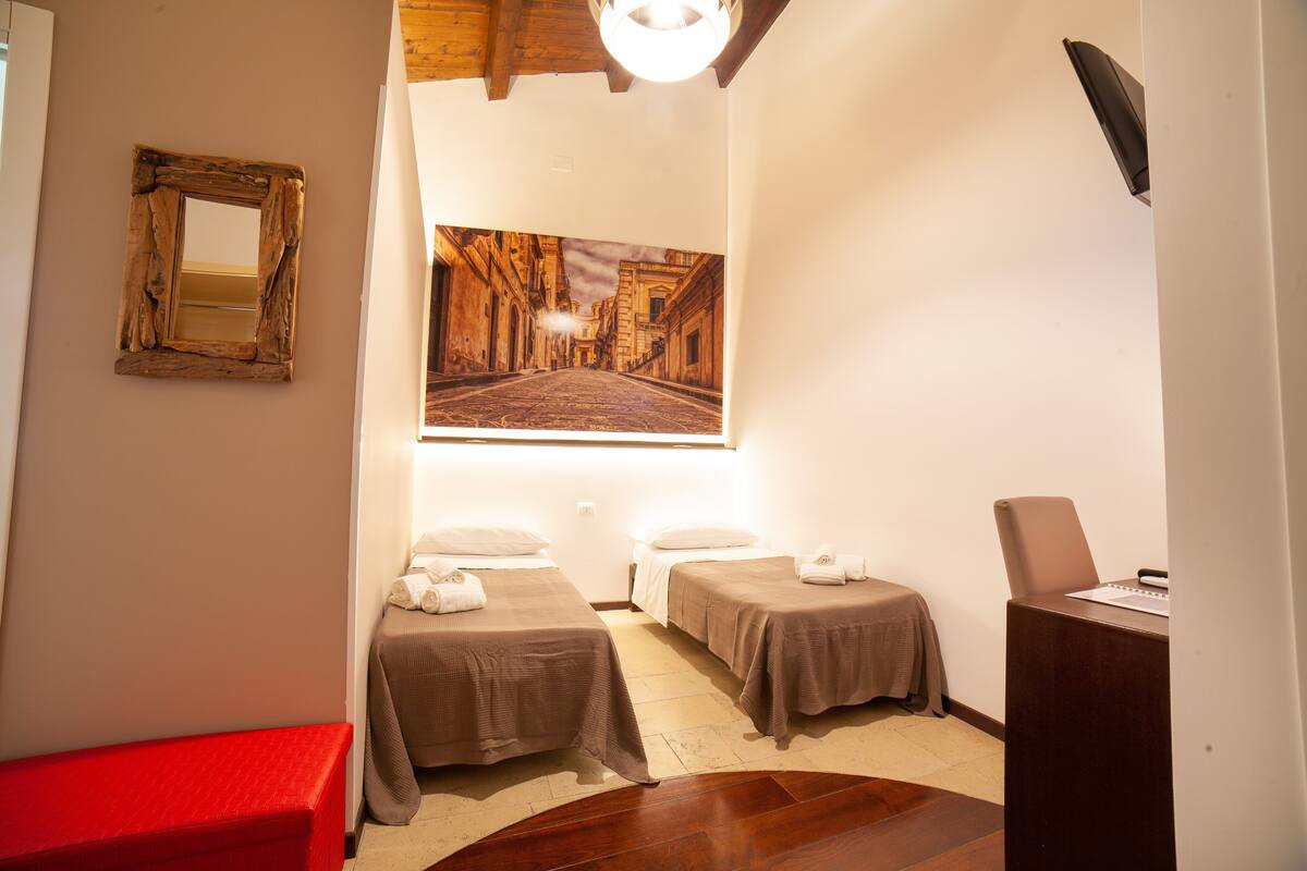 A hotel room with two single beds, a red bench, a desk with a chair, and a large framed picture on the wall.