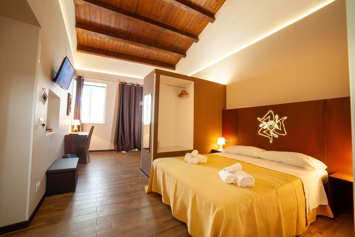 A hotel room with a wooden ceiling, a bed with a yellow bedspread, a bedside table with a lamp, and a TV mounted on the wall.