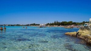 Une mer d’un bleu profond, parsemée de rochers, avec quelques personnes dans l’eau et des bâtiments qui bordent la côte.