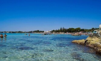 Mar azul cristalina con afloramientos rocosos, algunas personas en el agua y edificios a lo largo de la costa.
