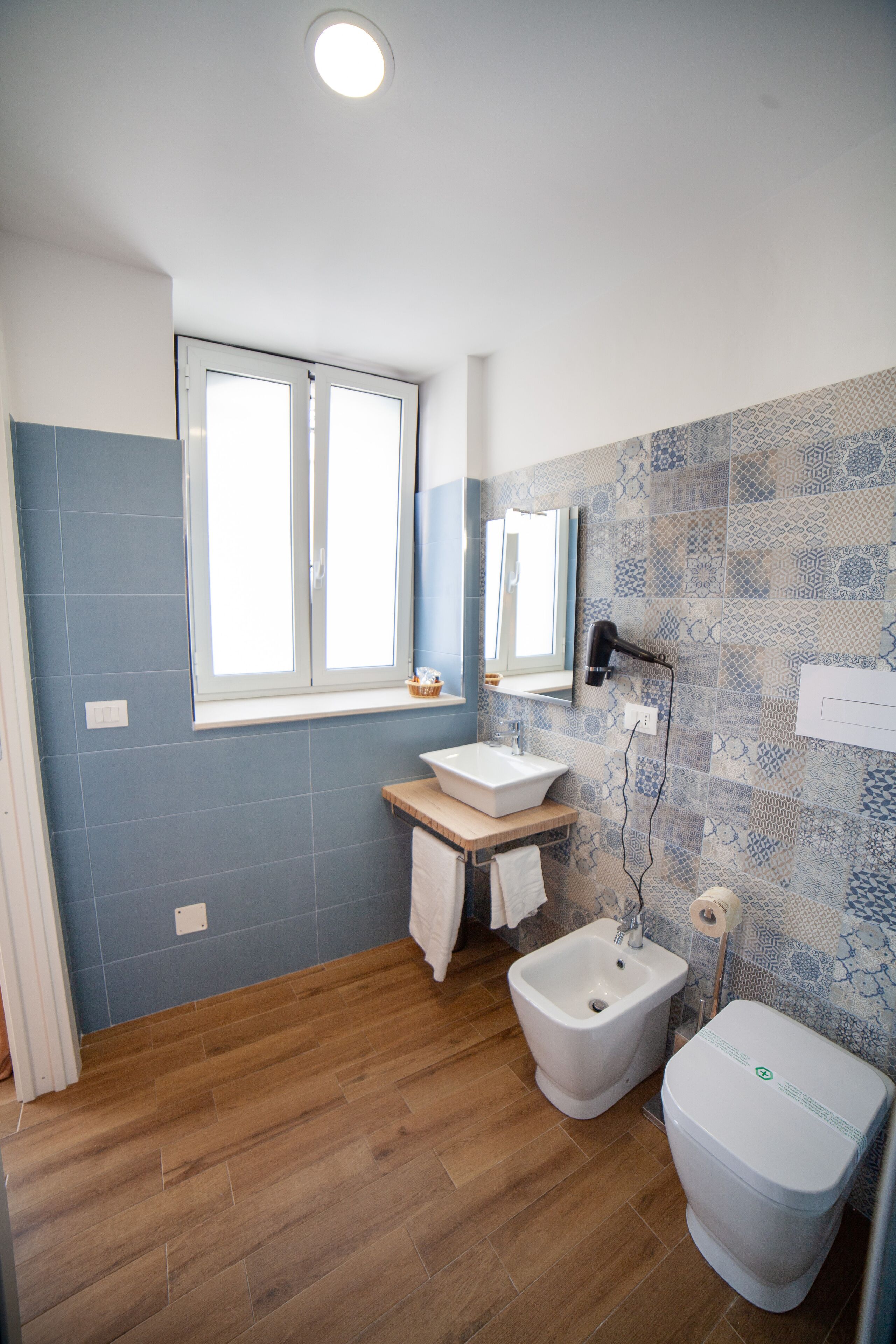 Een badkamer met een houten vloer, een wastafel, een toiletbril en een toilet. Er is een raam boven de wastafel en een spiegel aan de muur.