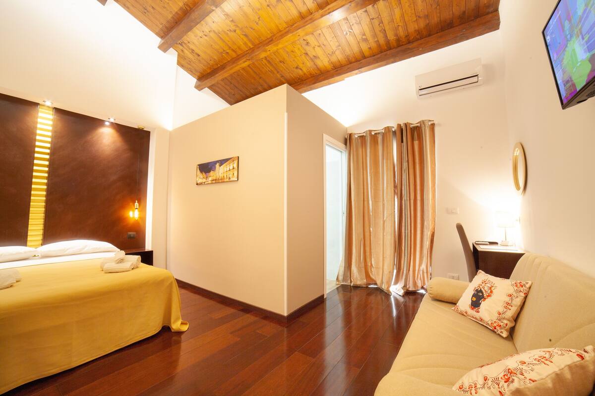 A hotel room with a bed, a sofa, wooden flooring, a painting, and an air conditioning unit.