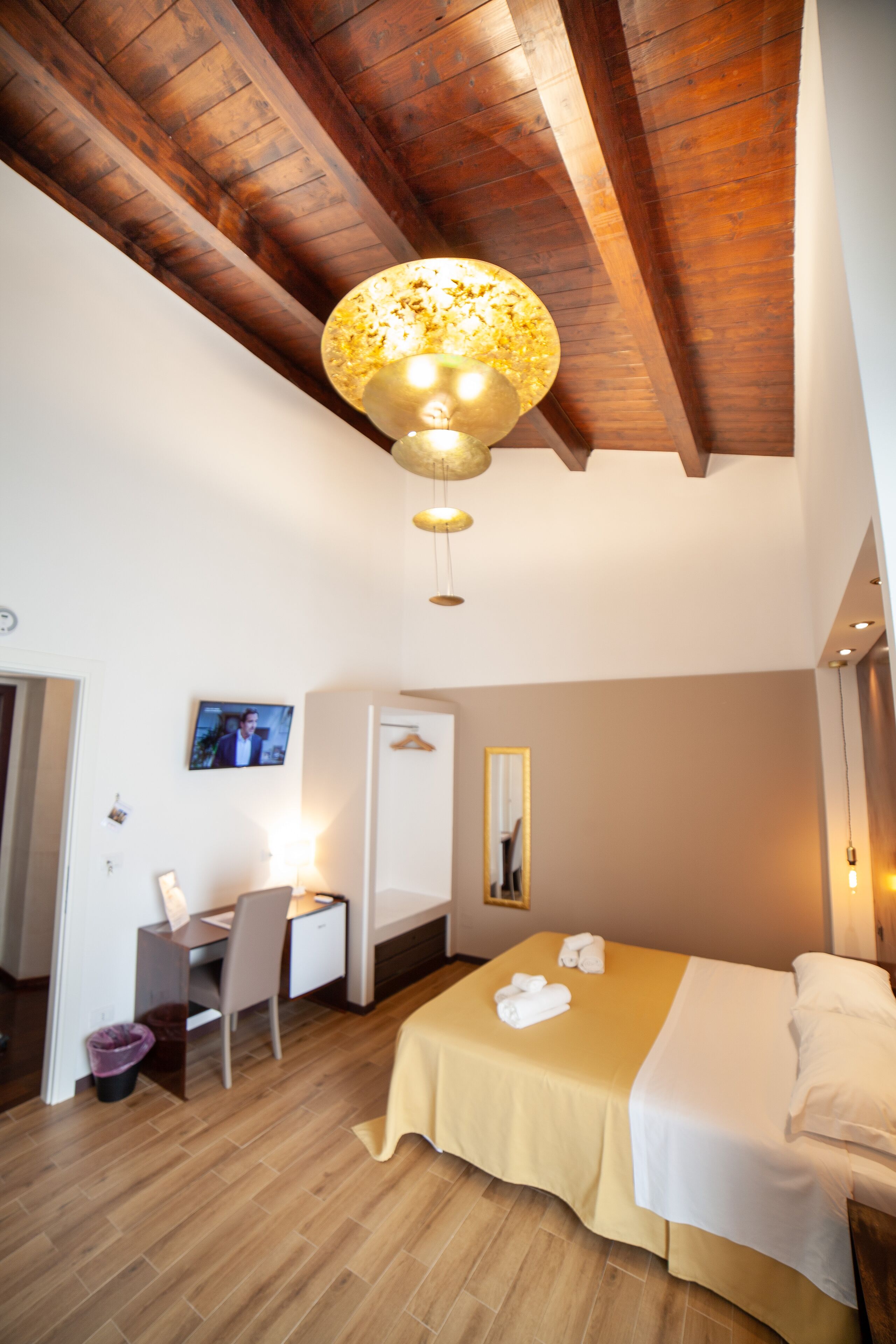 A hotel room with a wooden ceiling, a bed with a yellow bedspread, a desk with a chair, and a television on the wall.