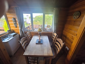 Ein Holzinnenraum mit einem Esstisch, Stühlen, einem Fenster mit Vorhängen und Blick ins Grüne.