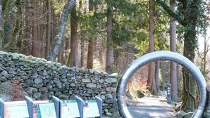 Un muro de piedra con un arco de metal, carteles informativos y un sendero arbolado.