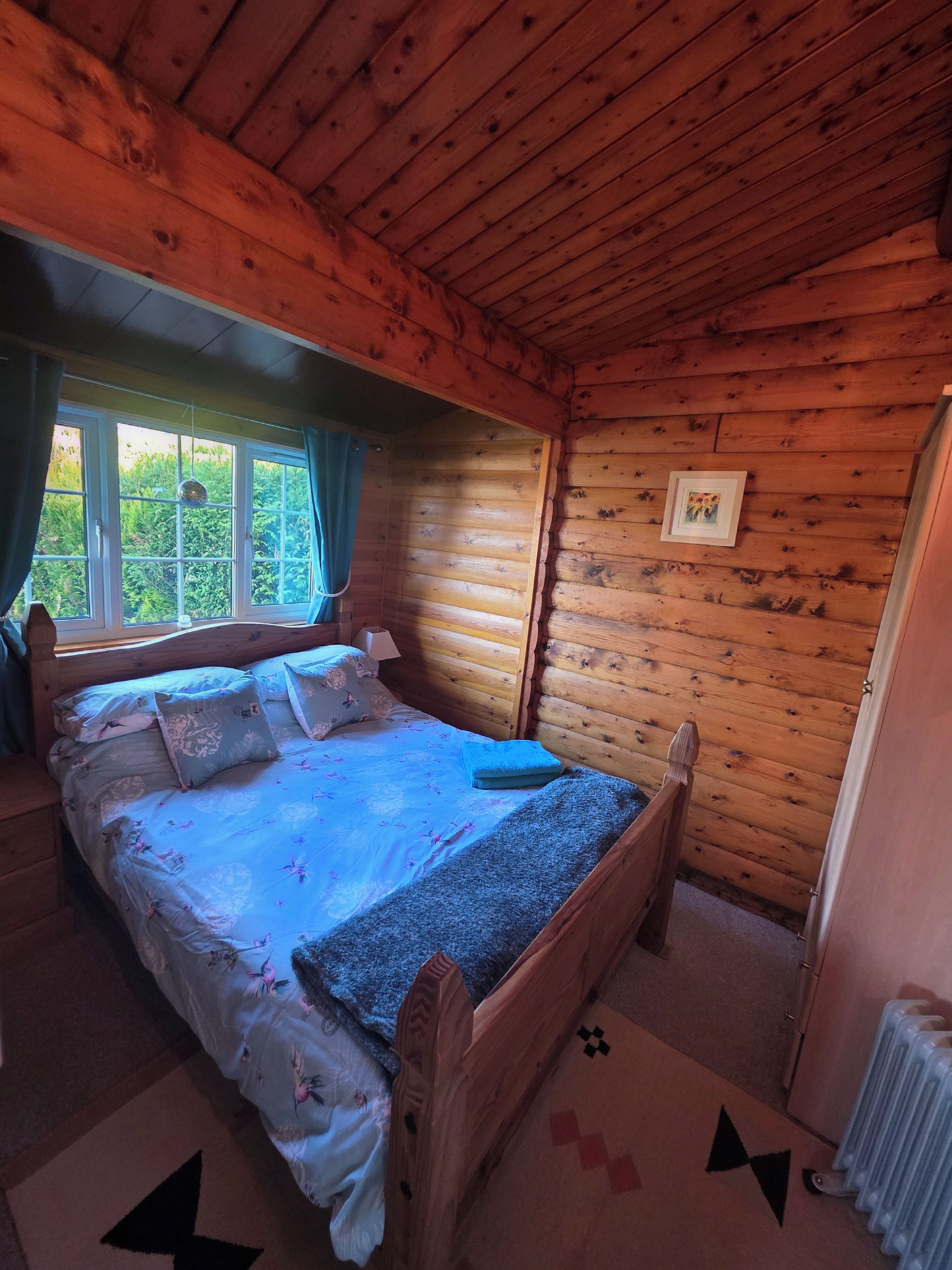 Quarto de cabana de madeira com cama, janela com cortinas, radiador e quadro pendurado na parede.