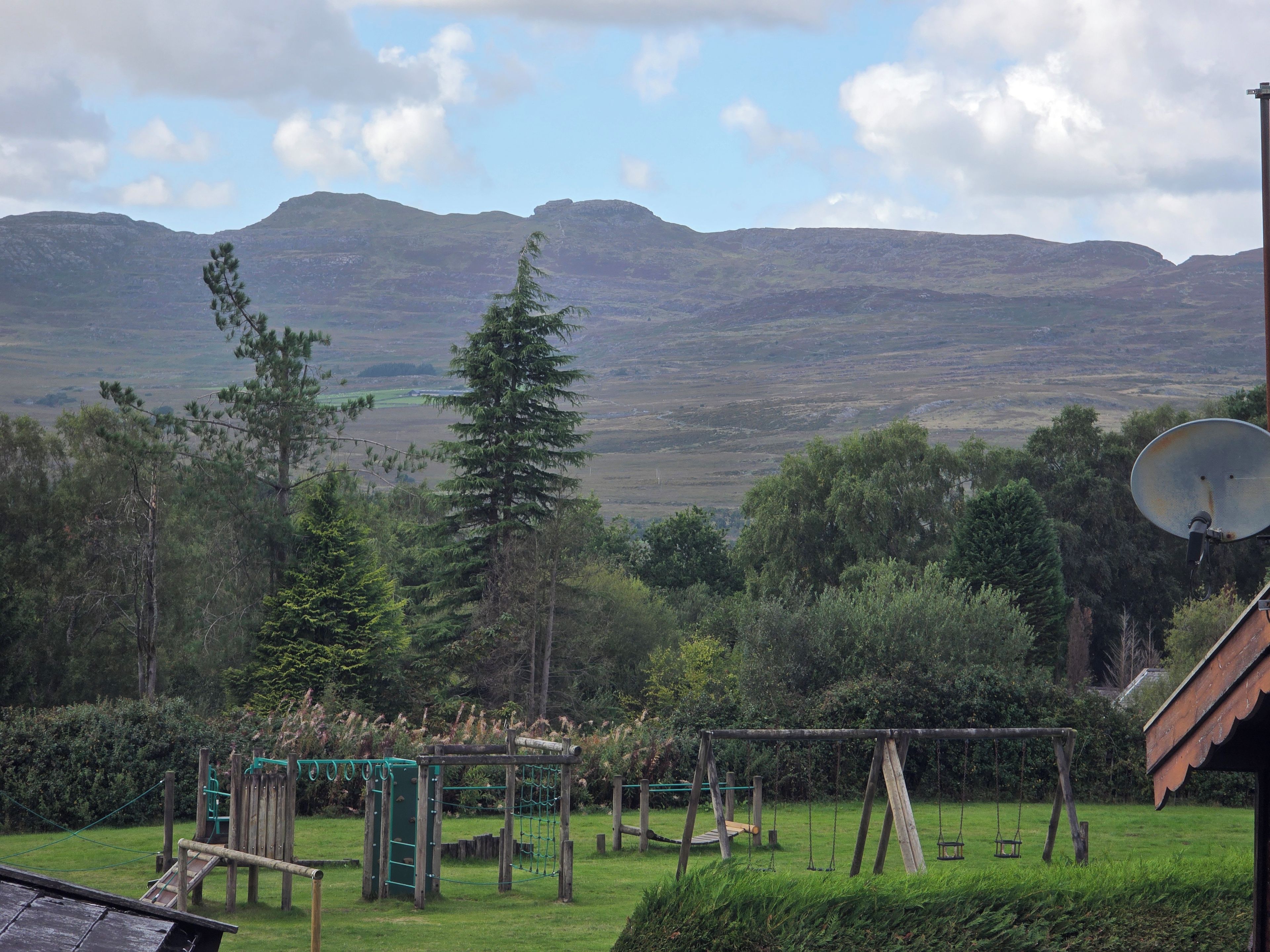 A playground with climbing structures and swings, surrounded by trees and a view of mountains.
