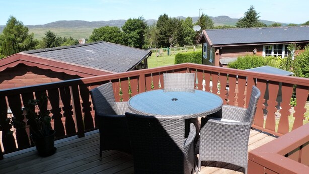Una terraza de madera con una mesa redonda y sillas de mimbre, con vistas a un paisaje pintoresco con colinas y árboles.