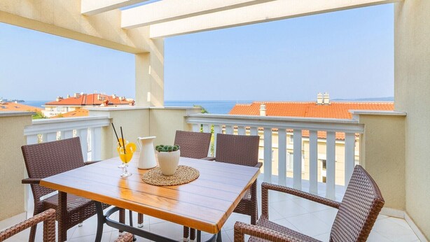 Ein Balkon mit Holztisch und Korbstühlen mit Blick auf eine Küstenstadt.