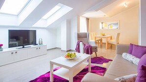 Ein modernes Wohnzimmer mit Dachfenstern, einem Flachbildfernseher, einem Essbereich und einem Sofa mit violetten Kissen.