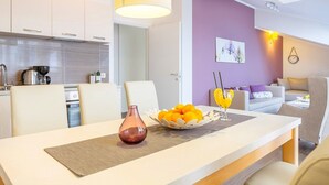 Eine moderne Küche mit weißen Schränken, einer Kaffeemaschine und einem Wasserkocher. Ein Essbereich mit Tisch für zwei Personen, inklusive einer Vase und einer Obstschale. Der Raum verfügt über eine violette Akzentwand und eine Wanduhr.
